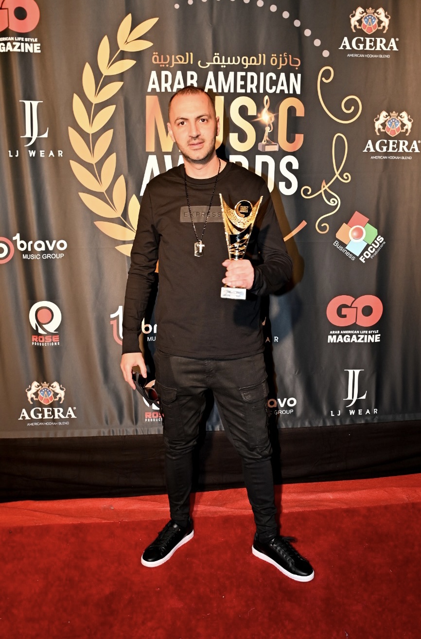 DJ holding an award at the Arab American Music Awards, standing in front of a branded backdrop, showcasing his recognition in the music industry. Top Rated DJ, Corporate DJ, Multi cultural DJ, Arabic DJ