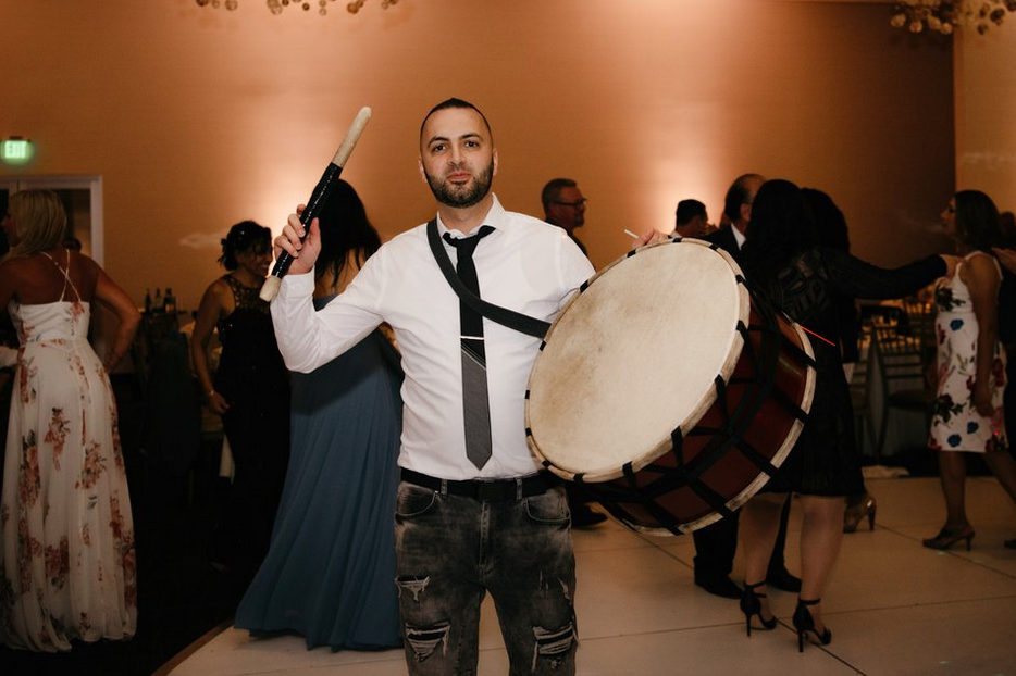 Man holding a large drum and stick, performing at a multicultural event, with guests dancing in the background, emphasizing DJ Franky Flow's Arabic music services. Best arabic Dj in California, Arabic wedding DJ